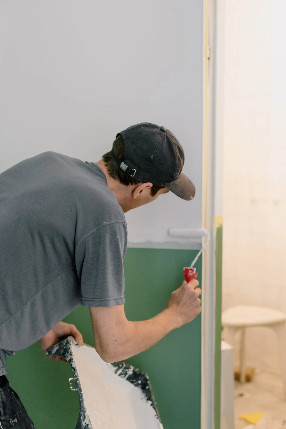 A man wearing a ball cap and a grey shirt rolling paint onto the top half of a two tone white-green wall. He is holding a paint tray with white paint in it.