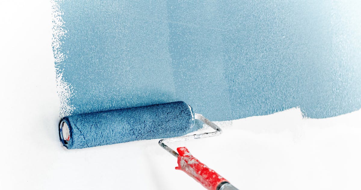 A paint roller on an extension handle, putting blue paint on a white interior wall.