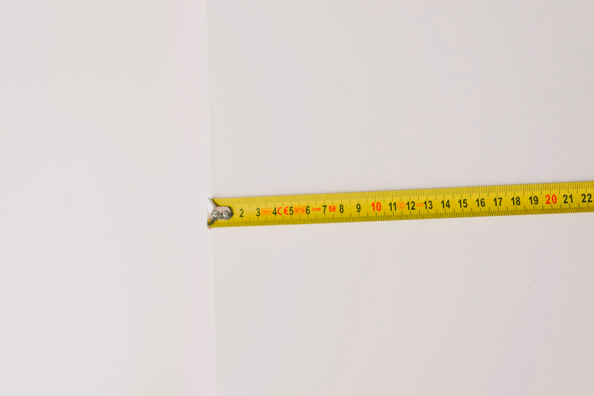 A yellow tape measure being held against the corner of a white wall, measuring a horizontal distance. The tape goes out of the image to the right 